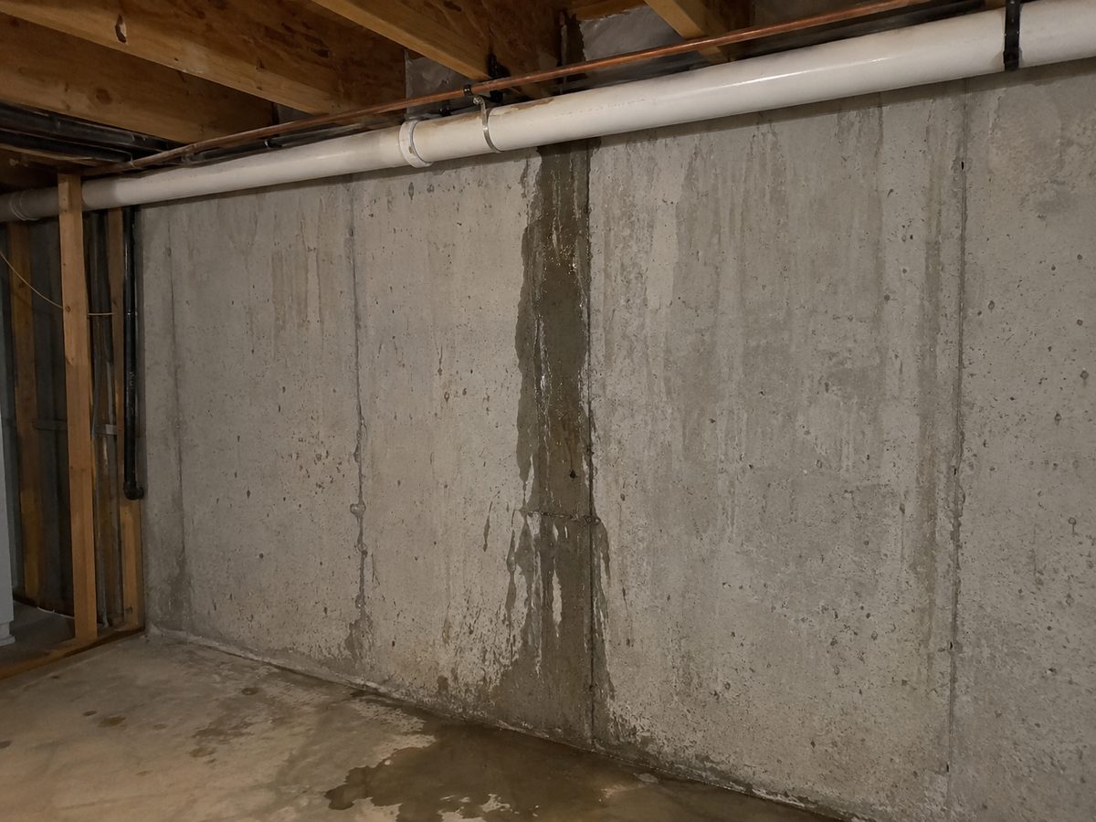 Dark wet streak running down an unfinished poured concrete basement wall from beneath a white PVC pipe and copper water line, with a puddle on the concrete floor