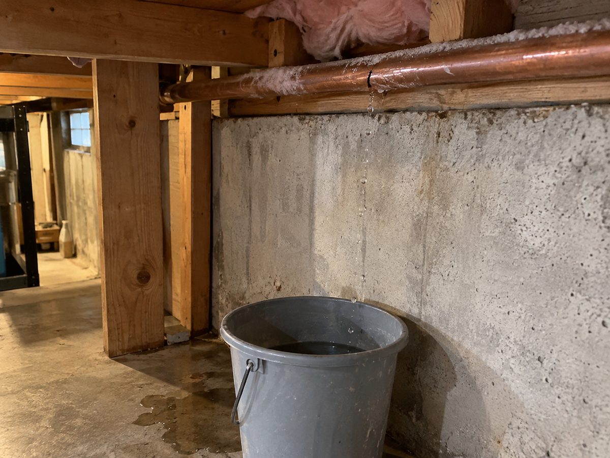 Frost-covered copper water pipe with a split, dripping into a gray bucket along an unfinished basement foundation wall