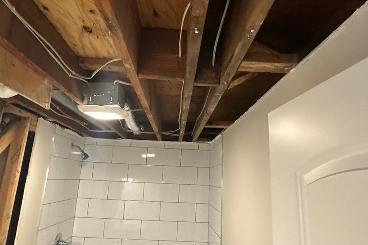 Bathroom with ceiling drywall removed above a tiled shower, exposing joists, wiring and subfloor after an upstairs leak