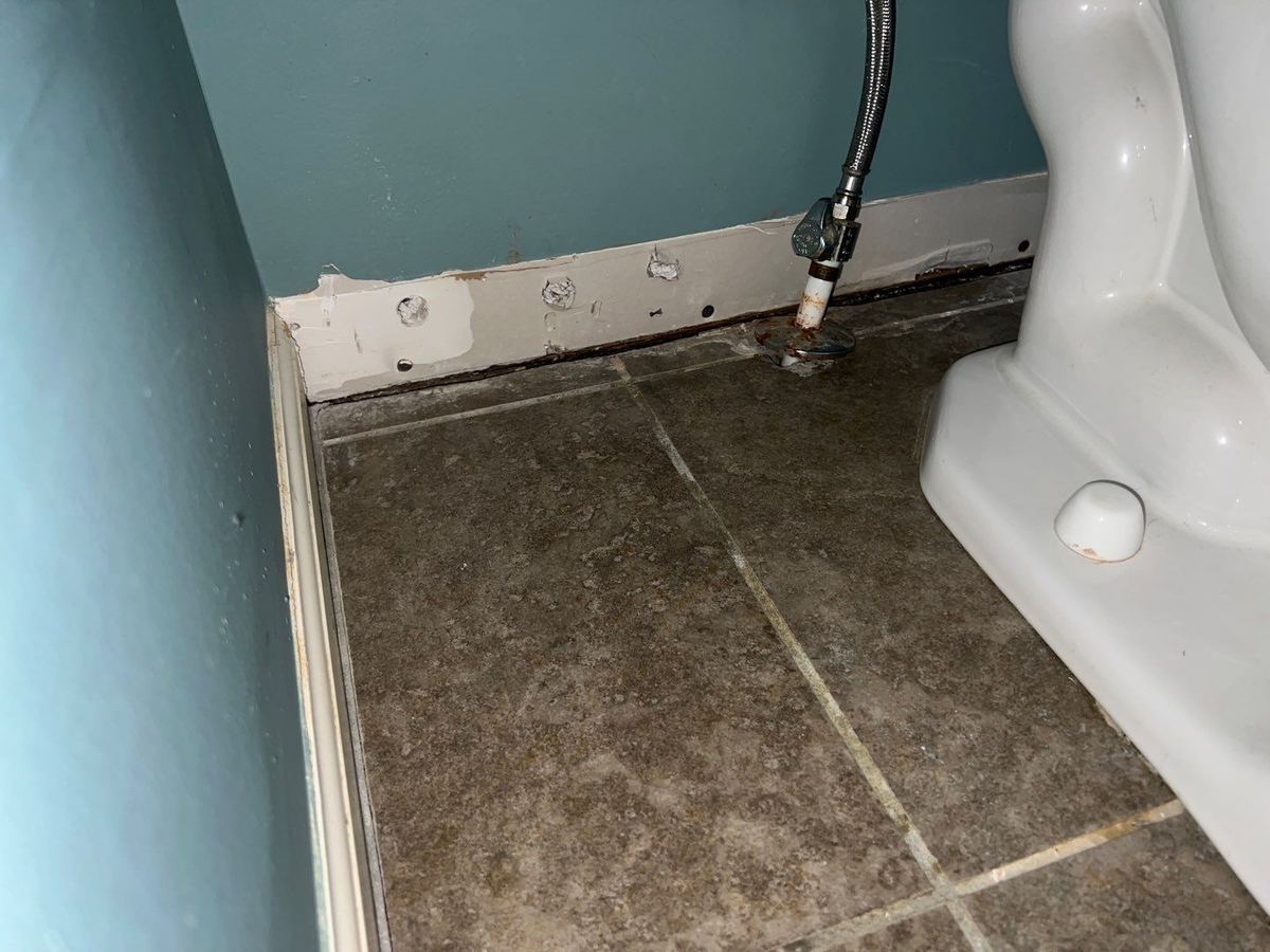 Baseboard removed and small drying holes drilled along the bottom of a bathroom wall after a supply line leak