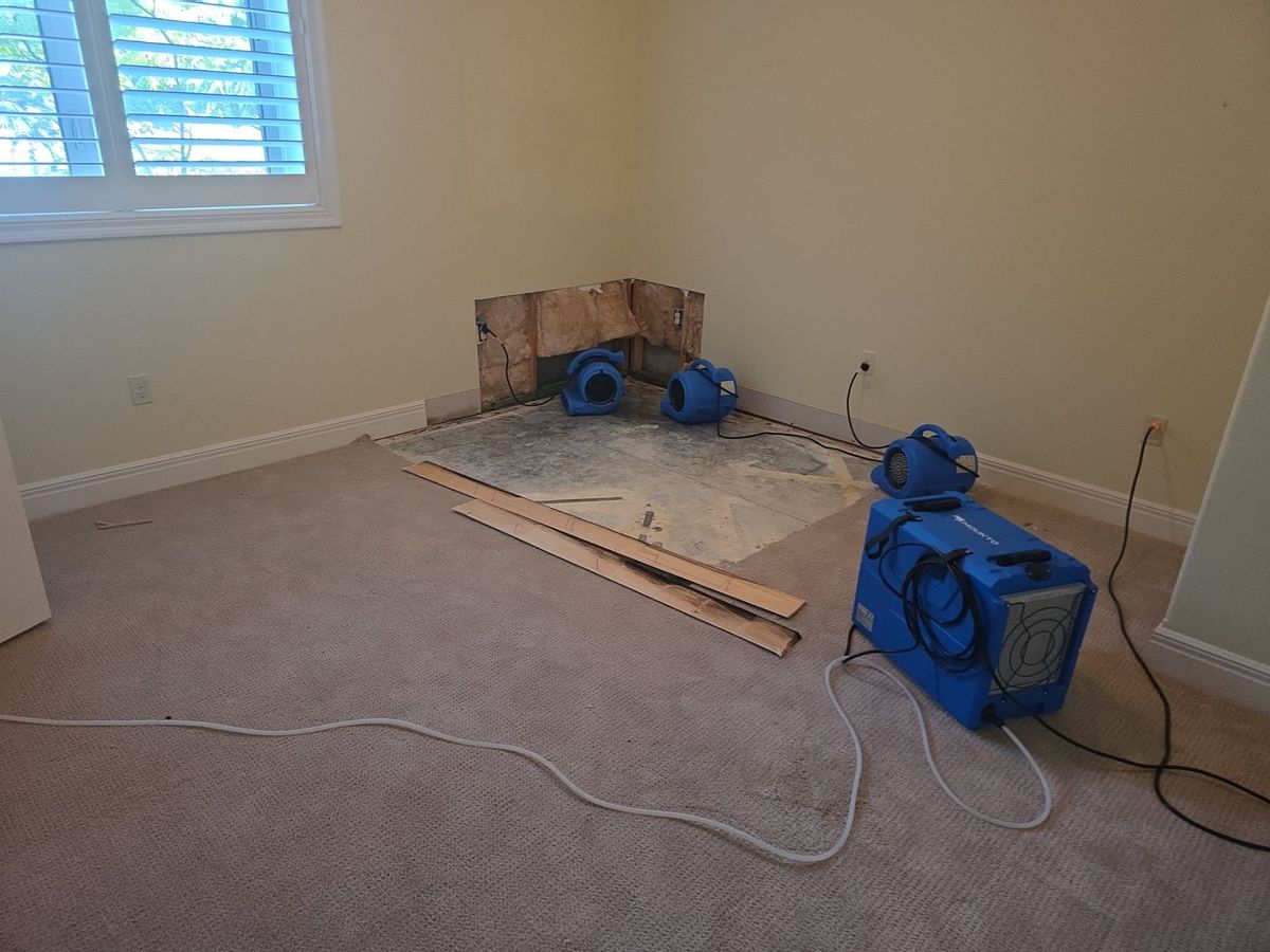 Bedroom corner with carpet pulled back, a section of lower drywall cut open, and three air movers and a dehumidifier drying the wall and subfloor